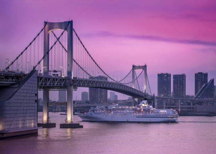 The Symphony, a luxury cruise ship, sailing under Rainbow Bridge as the sun sets. The sky is a vibrant pink.